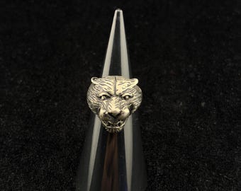 May include: A silver-toned metal ring featuring a detailed, three-dimensional tiger head design. The tiger face is sculpted with visible fur texture, pointed ears, and an open mouth showing teeth, centred on a smooth, polished metal band.