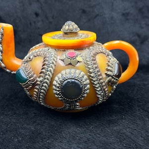 May include: A small, amber-colored teapot with intricate silver and gold embellishments. The teapot is decorated with colorful gemstones and has a traditional design.