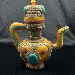 Op de afbeelding: Een sierlijke, barnsteenkleurige theepot met een decoratief kroonachtig deksel. De theepot heeft turquoise en rode accenten, met ingewikkeld metaalwerk en een gedetailleerde handgreep en tuit.