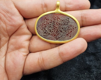 Pode incluir: Um pingente de ouro com uma pedra de cornalina vermelha com caligrafia árabe intrincada. O pingente tem forma oval e tem um aro de ouro.