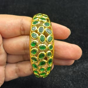 May include: A gold-toned bangle bracelet featuring an openwork lattice design. The surface is densely set with numerous small, oval-shaped, vibrant green gemstones arranged in a repeating pattern across the entire curved metal structure.