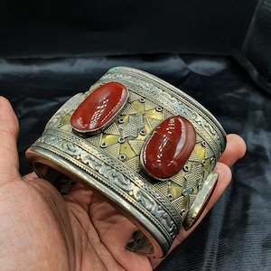 Pode incluir: Uma pulseira larga de metal em tons de prata e latão com padrões gravados intrincados e texturas geométricas. Duas grandes pedras ovais de cor vermelho-escura, lisas e brilhantes, estão cravadas na parte frontal da banda metálica.
