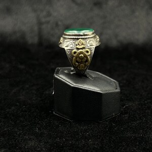 May include: A silver ring with a green stone and intricate gold detailing. The ring is on a black display stand.