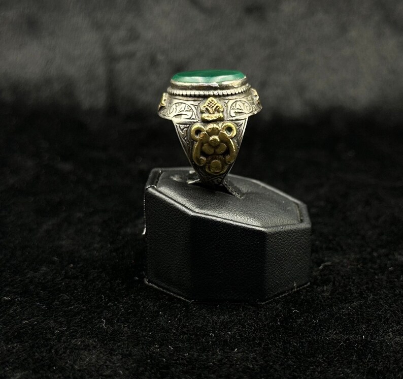 May include: A silver ring with a green stone and intricate gold detailing. The ring is on a black display stand.