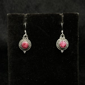 May include: A pair of silver earrings with a red gemstone set in a circular bezel. The earrings have a beaded border and a small drop.