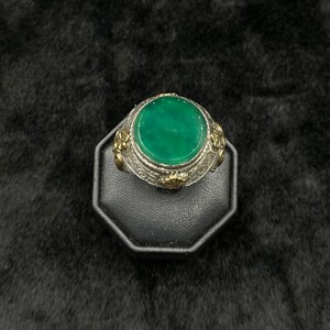 May include: A silver ring with a green gemstone set in a round bezel. The ring is decorated with floral designs and has a gold accent.