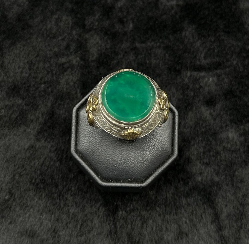 May include: A silver ring with a green gemstone set in a round bezel. The ring is decorated with floral designs and has a gold accent.