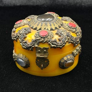 Op de afbeelding: Een ronde, gele sieradendoos met sierlijke metalen details. Het deksel heeft een grote centrale steen en is omgeven door rode en gouden accenten. De doos heeft een metalen sluiting en is waarschijnlijk vintage.