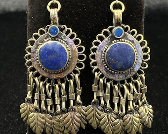 May include: A pair of ornate earrings featuring large, round, deep blue gemstones at the center. The earrings have a detailed, antique gold-colored metalwork design with dangling leaf-shaped charms. The earrings are displayed on a black surface.