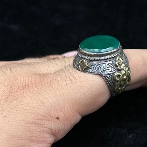 May include: A silver ring with an oval green stone set in a silver bezel. The ring is decorated with intricate floral designs and a gold accent.