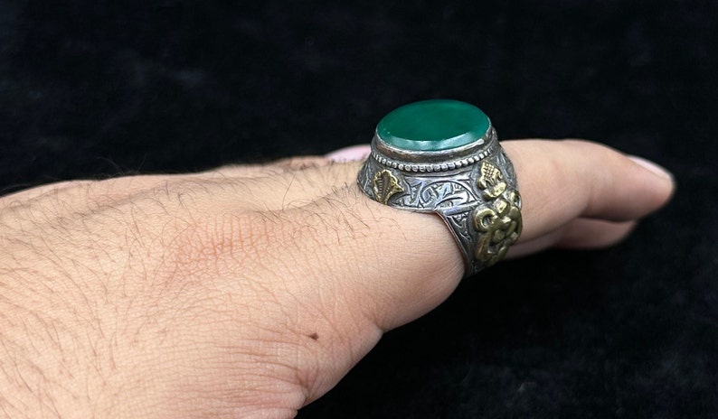May include: A silver ring with an oval green stone set in a silver bezel. The ring is decorated with intricate floral designs and a gold accent.