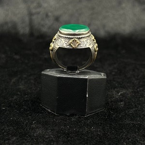 May include: A silver ring with a green stone set in a bezel setting. The ring is decorated with intricate floral designs and has a gold accent.