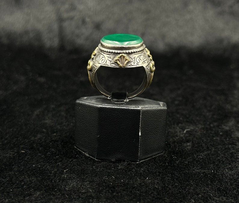 May include: A silver ring with a green stone set in a bezel setting. The ring is decorated with intricate floral designs and has a gold accent.