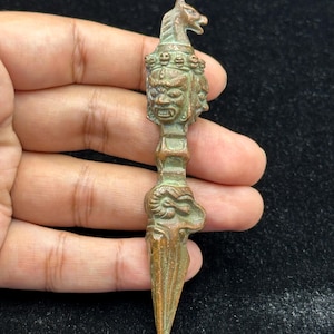 May include: A bronze-colored, ornate dagger with a horse head at the top and a detailed face design. The dagger has a pointed tip and is held in a hand against a dark background. The dagger's design includes multiple faces and intricate details.
