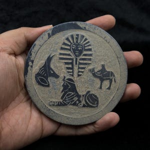 May include: A circular, grey stone coaster featuring black etched Egyptian motifs. The design includes a pharaoh's head, a sphinx, a camel, and a jackal. The coaster is held in a hand against a dark backdrop.