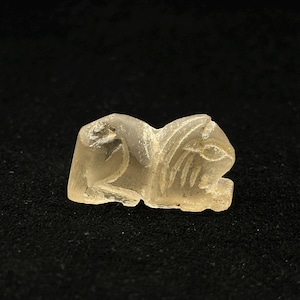 May include: A small, translucent, pale yellow carved stone figurine depicting a crouching animal in profile. The carving features etched lines representing the creature's facial features, mane, and limbs, with a smooth, polished surface texture.