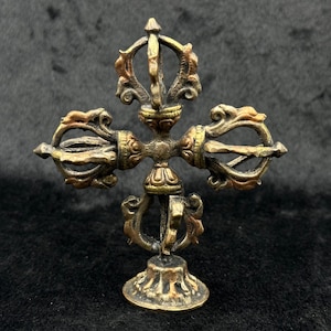 May include: A gold and copper colored metal sculpture of a Tibetan prayer wheel with intricate details. The prayer wheel is on a round base and has four arms with a central spire.