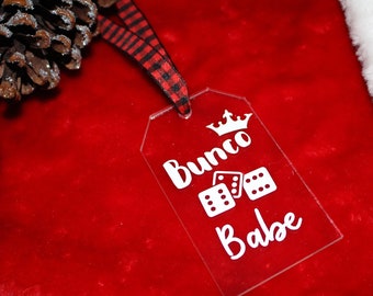 May include: A clear acrylic tag with the words "Bunco Babe" and dice illustrations in white. The tag is attached to a red and black plaid ribbon, set against a red velvet background with pine cones and a white fluffy edge.