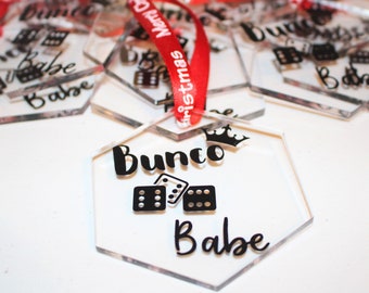 May include: Clear acrylic hexagon ornaments with black text and dice graphics. The ornaments have a red ribbon with white text that says "Merry Christmas". The text on the ornaments says "Bunco Babe".