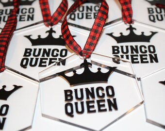 May include: Clear acrylic hexagon-shaped ornaments with the text "BUNCO QUEEN" in bold black letters, topped with a black crown graphic. Each ornament has a red and black plaid ribbon for hanging. The ornaments are designed for a Bunco game.