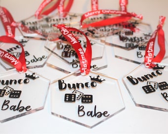 May include: Clear acrylic hexagon ornaments with black text that reads "Bunco Babe" and dice graphics. Each ornament has a red ribbon with "Merry Christmas" printed on it. A festive holiday decoration.