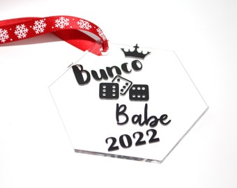 May include: A clear, hexagonal ornament with the words "Bunco Babe 2022" in black script, along with dice and a crown graphic. A red ribbon with white snowflakes is attached for hanging. A festive decoration.