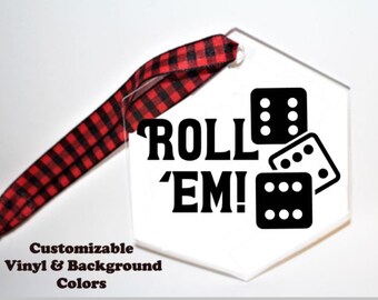 May include: A clear, hexagonal ornament with the words "ROLL 'EM!" and three dice with black dots. A red and black plaid ribbon is attached for hanging. The text "Customizable Vinyl & Background Colors" is also present.