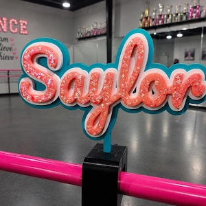 Op de afbeelding: Een gepersonaliseerd naambord met de naam Saylor in een cursief lettertype. De letters zijn glinsterend koraal-oranje, gelaagd over een witte omlijning en een effen blauwgroene achtergrond. Het bord is bevestigd aan een roze balletbarre.