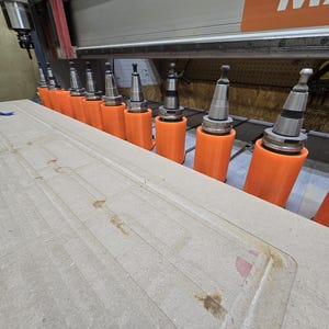 May include: Close-up of a CNC machine with multiple drill bits attached to orange holders. The machine is cutting a design into a piece of wood.
