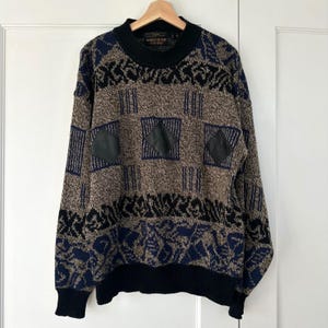 May include: A vintage sweater with a brown, black, and navy blue geometric pattern. The sweater features black leather patches and a black ribbed collar, cuffs, and hem. The sweater is hanging on a wooden hanger.