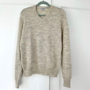 May include: A beige V-neck sweater with a textured, herringbone pattern. The sweater is made of a soft material and has ribbed cuffs and hem. The sweater is hanging on a green hanger against a white door.