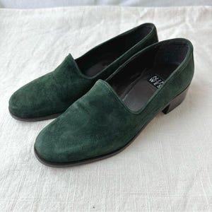 May include: A pair of dark green suede slip-on shoes. The shoes have a rounded toe and a low heel. The brand name "Nine West" is visible on the insole of one shoe. The shoes are set against a white background.