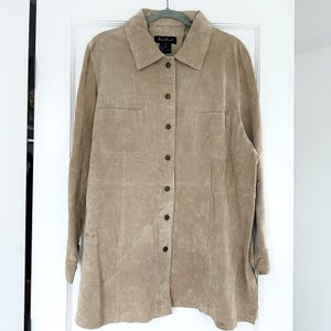 May include: A tan suede button-down shirt with two front pockets and a collared neckline. The long-sleeved shirt has a classic design with a button closure and is hanging on a hanger. The shirt is a neutral color.