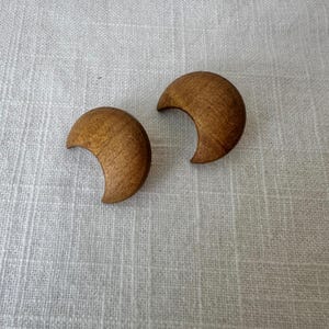 May include: A pair of crescent moon-shaped earrings crafted from smooth, light brown wood. Each piece features a distinct natural wood grain pattern, showcasing a curved, solid design that mimics the shape of a waxing or waning moon.