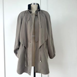 May include: A taupe-colored trench coat with a black collar and a zip-up front. The coat has a drawstring waist, long sleeves, and a layered design. The coat is displayed on a mannequin.