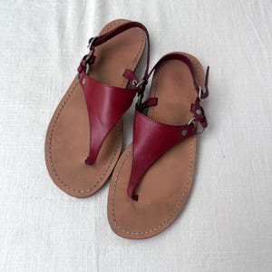 May include: A pair of red leather sandals with a thong-style design. The sandals have brown soles and adjustable ankle straps with silver-colored metal hardware. The edges are stitched with a light-colored thread.