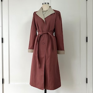 May include: A long, dusty rose trench coat with a contrasting tan collar and cuffs. The coat features a matching belt, a button closure, and a classic design. The coat is displayed on a mannequin.