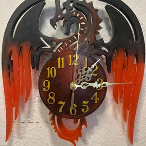 May include: A dragon-themed wall clock with a brown face and gold hands. The clock features a dragon design with black wings and orange accents. The numbers are gold. The clock is mounted on a white wall.