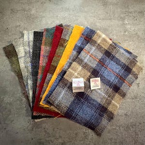 May include: A collection of colorful Harris Tweed fabric swatches, showcasing a variety of plaid and solid patterns. The swatches include shades of blue, red, yellow, and green, with two small labels visible. The fabrics are arranged in a fan shape.