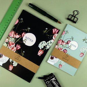 Könnte beinhalten: Zwei Blumen-Notizbücher mit braunen Papierumschlägen und dem Text "NOTES:" und "FUR DICH GEMACHT". Die Notizbücher haben ein schwarz-weißes Blumenmuster mit rosa Blüten.
