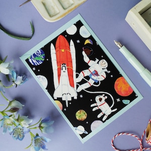 Peut inclure: Carte de vœux sur le thème de l'espace. La carte présente une fusée rouge et blanche, un astronaute et un chien en combinaison spatiale, entourés de planètes et d'étoiles sur fond noir. La carte a une bordure bleu clair.