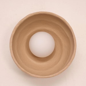 Op de afbeelding: Een lichtbruine, getextureerde kom met een witte bol in het midden. De kom heeft een afgeronde vorm met een enigszins ongelijke rand. De bol is glad en ondoorzichtig, wat contrasteert met de ruwe textuur van de kom.