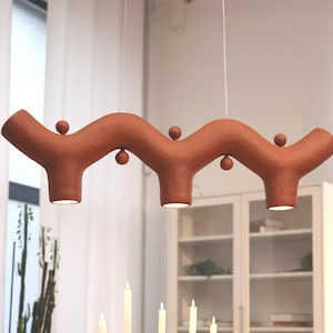 May include: A terracotta-colored pendant light fixture with a wavy, branch-like structure featuring three downward-facing lights and small spherical accents, suspended above a round wooden dining table with a metal candelabra and place settings.