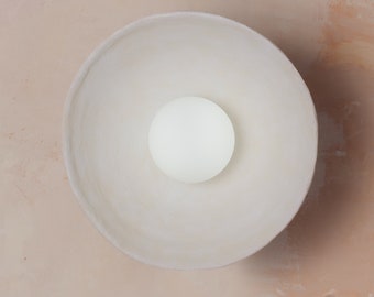 May include: A circular, off-white ceramic bowl with a smooth, matte finish and an irregular, organic rim. Centered inside the bowl is a single, solid white sphere, creating a stark contrast against the muted, light-colored interior surface.