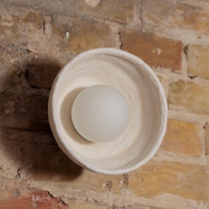 May include: A white ceramic light fitting with a round, bowl-shaped design. The light fitting is mounted on a brick wall and has a white light bulb inside.