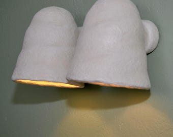 Op de afbeelding: Een witte, getextureerde wandlamp met twee klokvormige armaturen. De lamp is bevestigd aan een lichtgroene muur. De binnenkant van de armaturen gloeit met een warm, geel licht en werpt een zachte schaduw eronder.