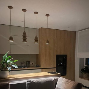 Könnte beinhalten: Ein modernes Wohnzimmer mit einem braunen Sofa und einer Küche im Hintergrund. Die Küche hat eine Arbeitsplatte aus Holz und weiße Schränke. Fünf Pendelleuchten hängen von der Decke.