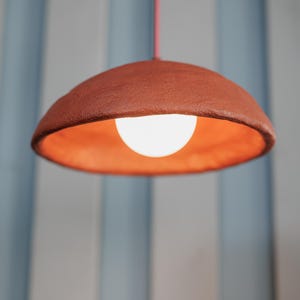 Pode incluir: Uma lumin&aacute;ria pendente cor de terracota com uma sombra redonda em forma de tigela. O interior da sombra &eacute; laranja e uma l&acirc;mpada branca brilhante &eacute; vis&iacute;vel. A luz pende de um fino cabo vermelho contra um fundo de listras verticais azuis e brancas.