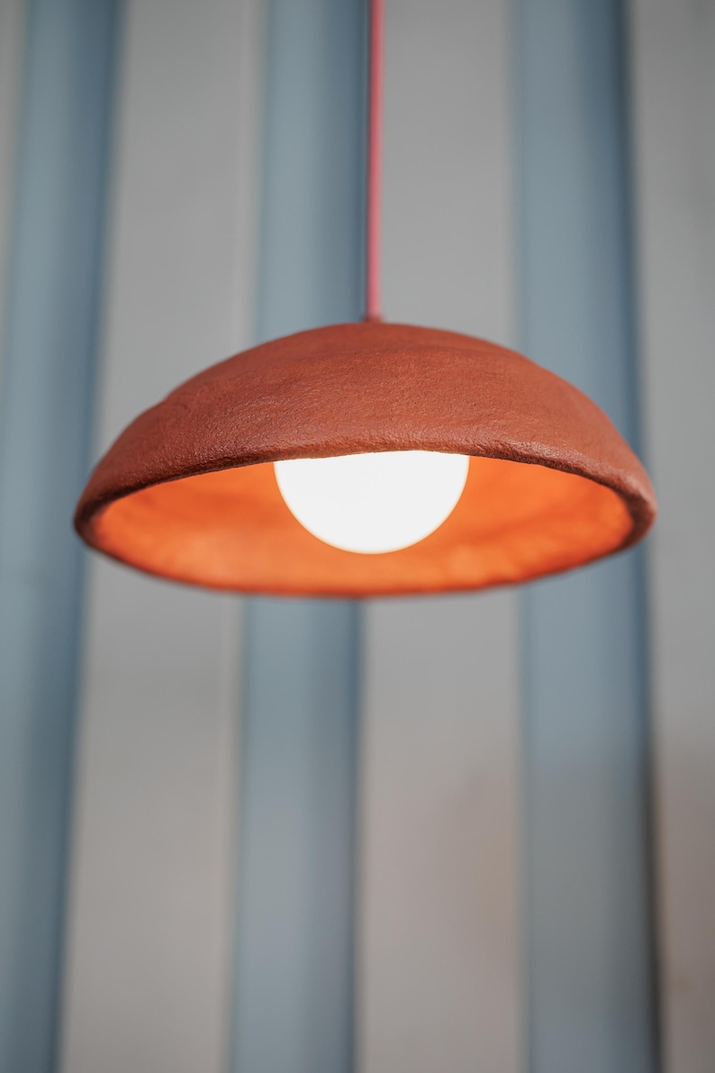 Pode incluir: Uma lumin&aacute;ria pendente cor de terracota com uma sombra redonda em forma de tigela. O interior da sombra &eacute; laranja e uma l&acirc;mpada branca brilhante &eacute; vis&iacute;vel. A luz pende de um fino cabo vermelho contra um fundo de listras verticais azuis e brancas.