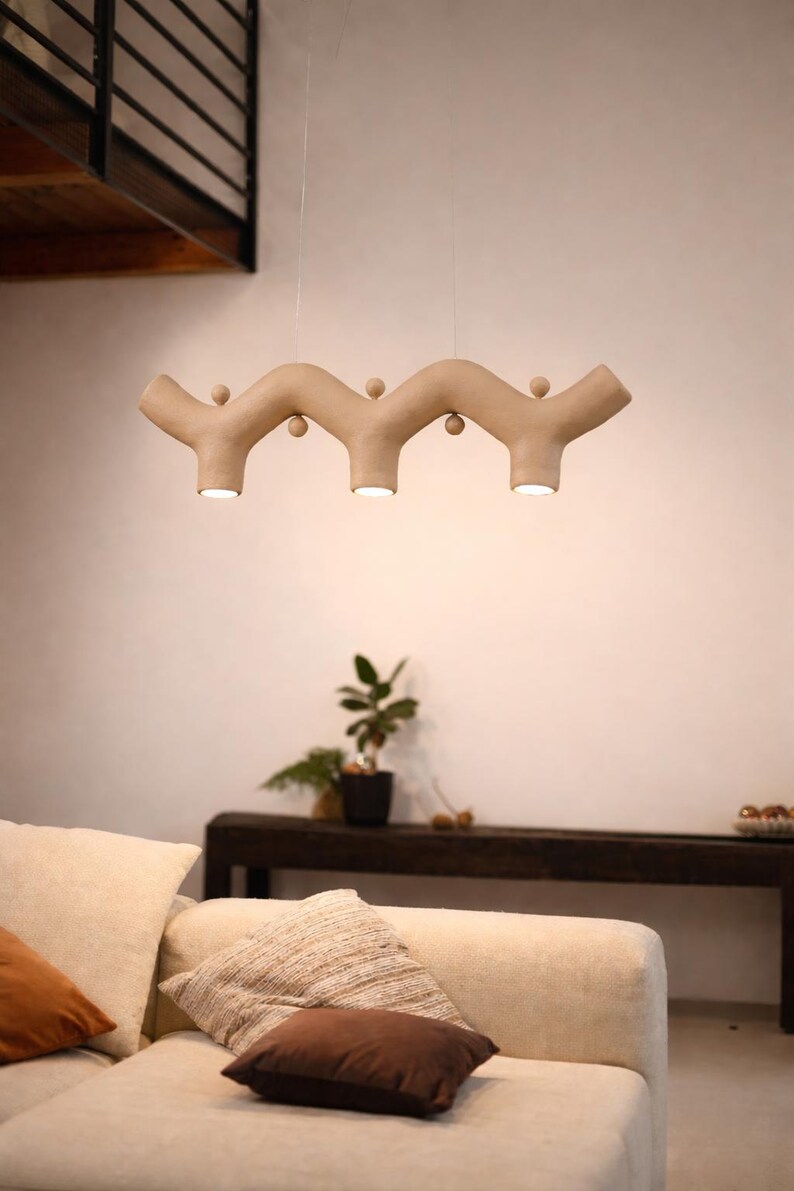 May include: A beige, wavy, horizontal pendant light fixture suspended by thin wires. The fixture features three downward-facing circular light sources and small spherical decorative accents positioned along the top and bottom curves of the structure.